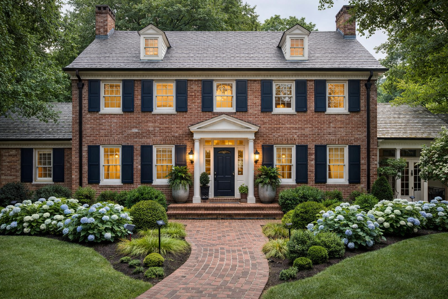Restored Maryland Colonial home exterior with classic architecture, white siding, stone accents, warm interior lighting, manicured landscaping, and a refined front entry at twilight.